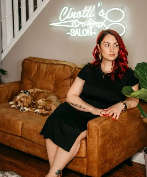 Sara on a cognac leather couch beside Chloe, a sleeping cockapoo, under a neon Cinotti's Salon sign
