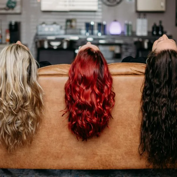 Three women lying back on a leather couch, their long blonde, red and dark brown curls hanging over the back