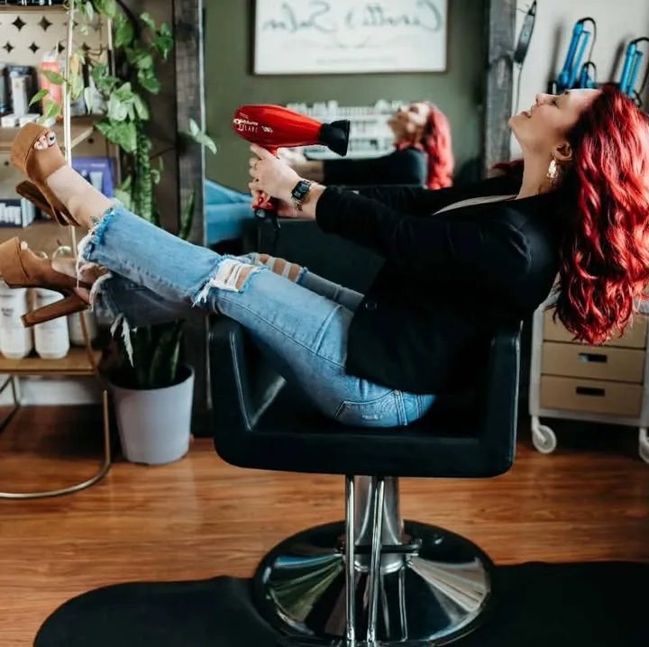 Sara, in a black jacket and ripped jeans, leans back in a styling chair with her feet up, holding a red hair dryer