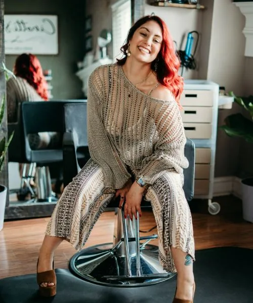 Sara, smiling, sitting on a styling chair in a knit sweater